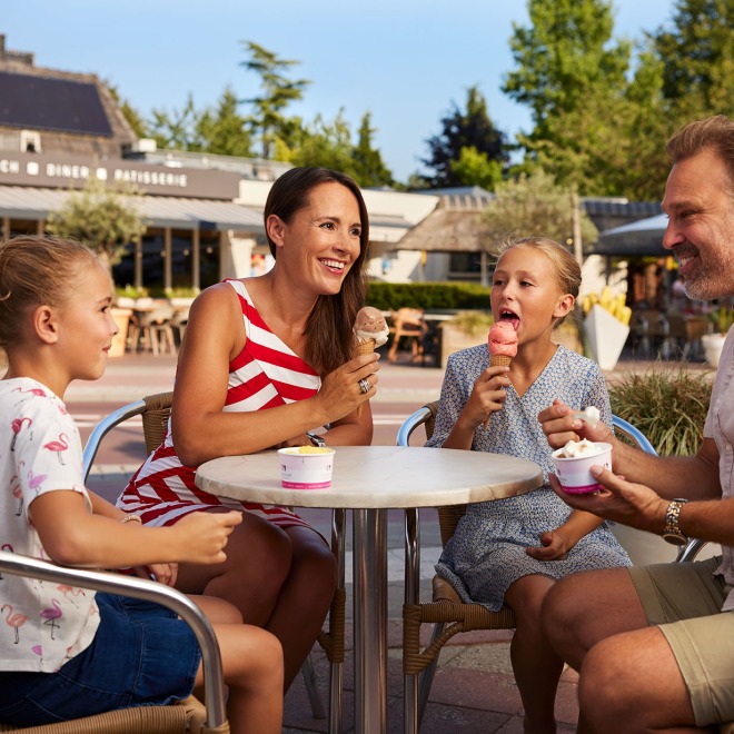 Gezin met twee kinderen eet samen ijsjes aan een buitentafel bij een restaurant op een zonnige dag.