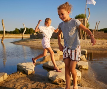 Twee kinderen lopen vrolijk over stenen in een ondiepe waterpartij tijdens een zonnige dag in de natuur.