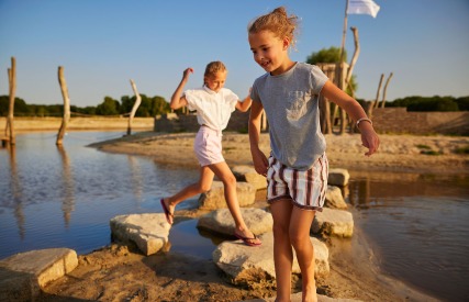 Twee kinderen lopen vrolijk over stenen in een ondiepe waterpartij tijdens een zonnige dag in de natuur.