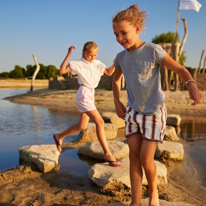 Twee kinderen lopen vrolijk over stenen in een ondiepe waterpartij tijdens een zonnige dag in de natuur.