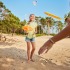 Twee kinderen spelen badminton op een zonnig strand met rackets en shuttle, omringd door bomen.