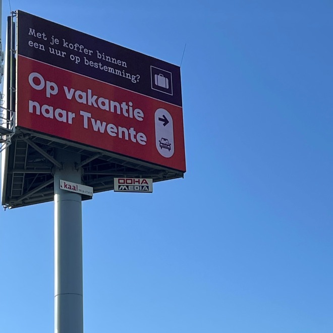 Reclamebord met de tekst 'Op vakantie naar Twente' en blauwe hemel op de achtergrond.