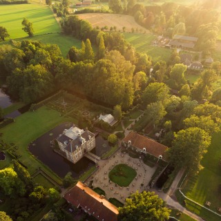Luchtfoto van een historisch kasteel met gracht, tuinen en groene velden bij zonsondergang.