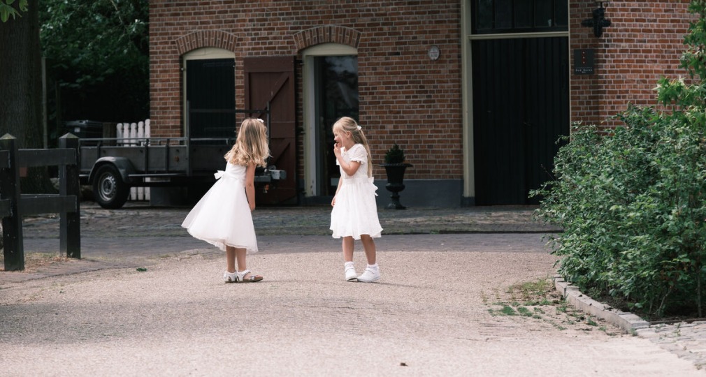 Twee jonge meisjes in witte jurken spelen buiten voor een traditioneel bakstenen huis op een zonnige dag.