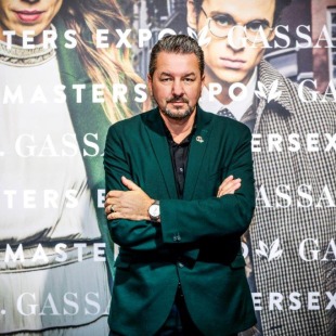 Man in groen colbert poseert met gekruiste armen voor Gassan Masters Expo achtergrond met modellen.