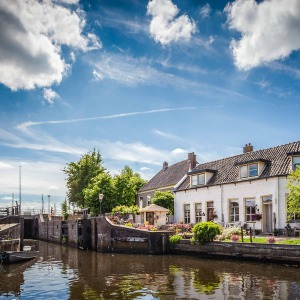 Rustiek Nederlands dorp met witte huizen aan een kanaal, bomen, bloemen, en wolkenluchten boven.