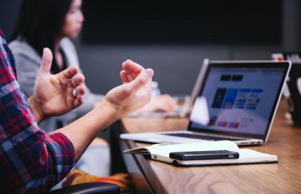 Close-up of a person gesturing while talking during a meeting, with a laptop and notebook on the table.