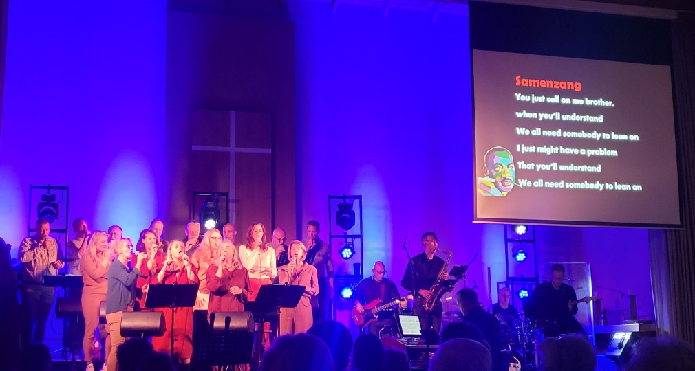 Muzikale uitvoering met koor en band in een kerk, songtekst van 'Lean on Me' geprojecteerd op scherm.