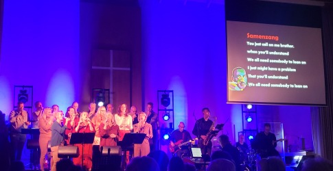 Muzikale uitvoering met koor en band in een kerk, songtekst van 'Lean on Me' geprojecteerd op scherm.