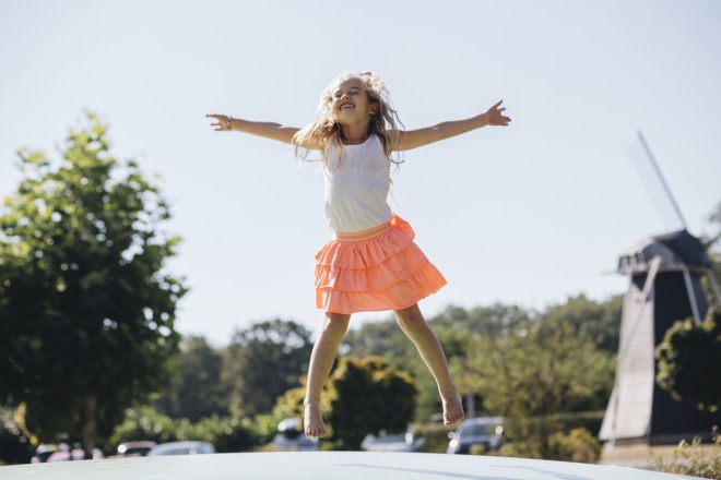 Een vrolijk meisje met een roze rok springt op een luchtkussen buiten, met bomen en een molen op de achtergrond.