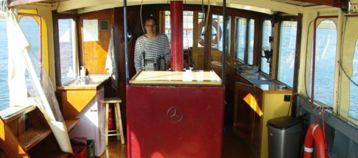 Interieur van een boot met houten banken, tafel, een stuurcabine, vrouw in gestreept shirt en uitzicht op water.