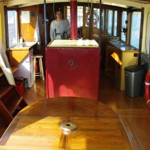 Interieur van een boot met houten banken, tafel, een stuurcabine, vrouw in gestreept shirt en uitzicht op water.