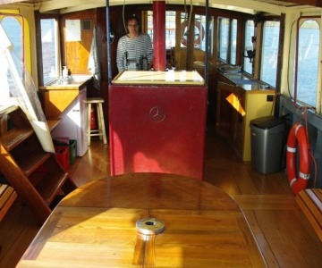 Interieur van een boot met houten banken, tafel, een stuurcabine, vrouw in gestreept shirt en uitzicht op water.