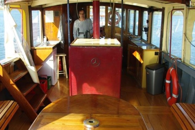 Interieur van een boot met houten banken, tafel, een stuurcabine, vrouw in gestreept shirt en uitzicht op water.