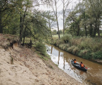 Twee mensen varen in een kano met reddingsvesten op een rustige rivier omgeven door bomen en zand.
