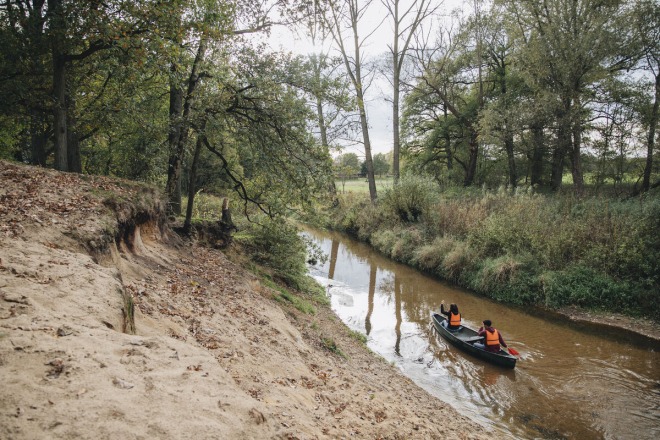 Twee mensen varen in een kano met reddingsvesten op een rustige rivier omgeven door bomen en zand.