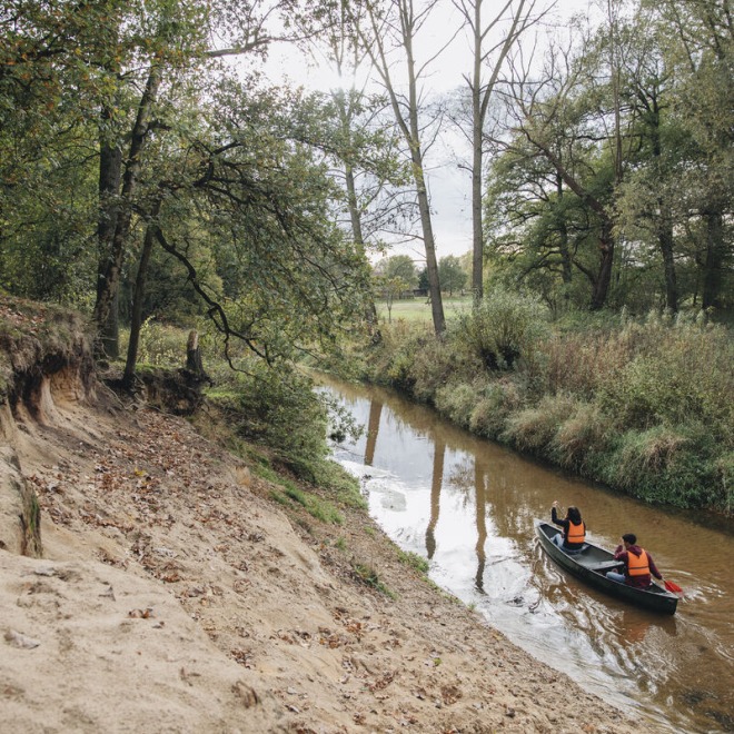 Twee mensen varen in een kano met reddingsvesten op een rustige rivier omgeven door bomen en zand.