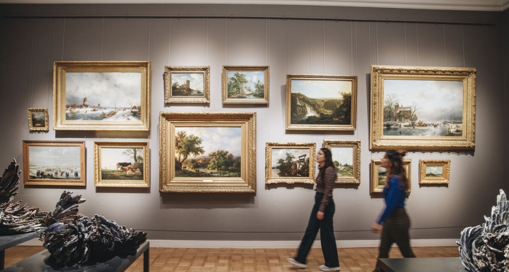 Twee mensen lopen langs een galerijmuur met verschillende schilderijen in een kunstmuseum.