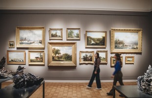 Zwei Personen gehen an einer Galerie mit gerahmten Gemälden in einem Kunstmuseum vorbei.