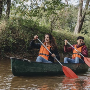 Twee mensen dragen reddingsvesten en kanoën samen op een rustige rivier omringd door bomen.