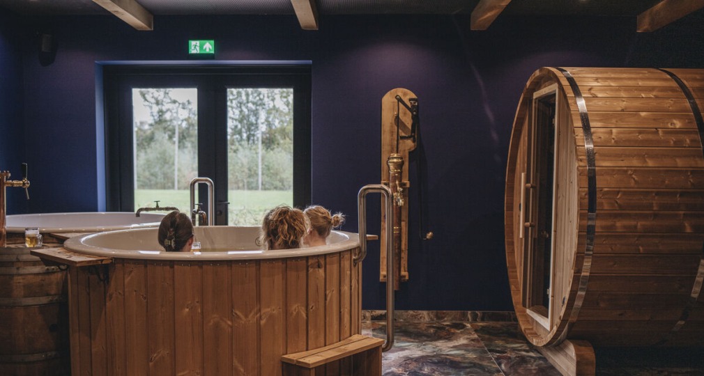 Binnenruimte met houten bubbelbad, drie mensen, tonvormige sauna en marmeren vloer, bij daglicht.