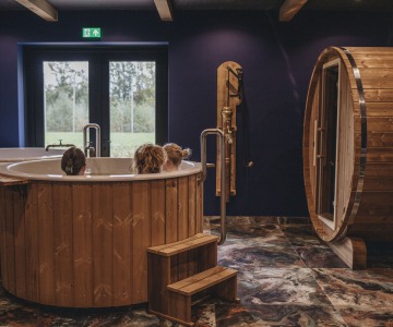 Binnenruimte met houten bubbelbad, drie mensen, tonvormige sauna en marmeren vloer, bij daglicht.