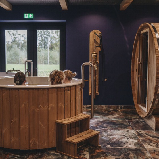 Binnenruimte met houten bubbelbad, drie mensen, tonvormige sauna en marmeren vloer, bij daglicht.