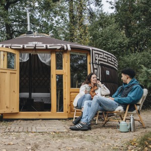 Twee mensen zitten buiten voor een traditionele yurt in het bos, pratend en genietend van de natuur.