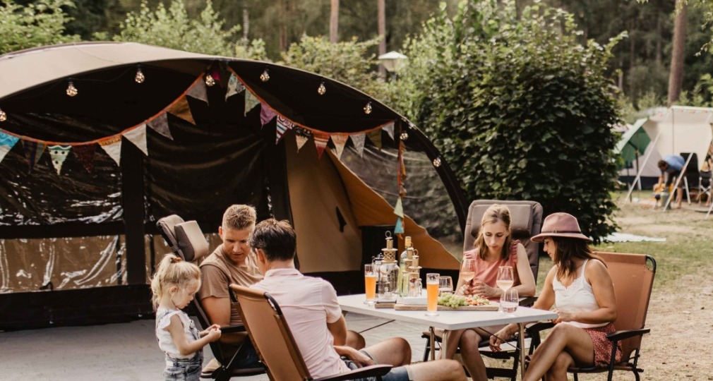 Gezin zit samen aan tafel voor een tent op een camping, omringd door bomen en andere kampeerders.