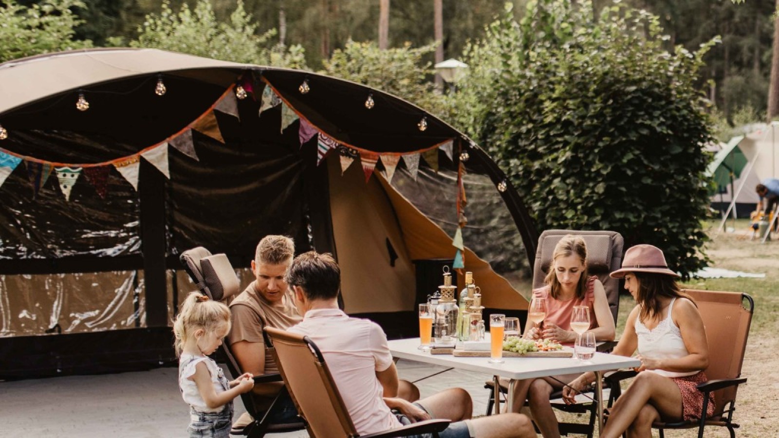 Gezin zit samen aan tafel voor een tent op een camping, omringd door bomen en andere kampeerders.