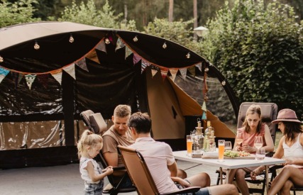 Gezin zit samen aan tafel voor een tent op een camping, omringd door bomen en andere kampeerders.
