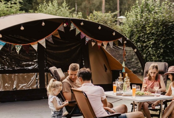 Gezin zit samen aan tafel voor een tent op een camping, omringd door bomen en andere kampeerders.