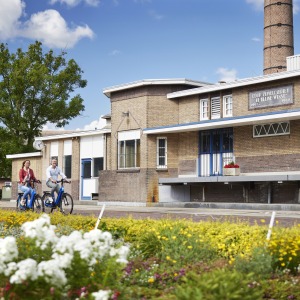 Twee mensen fietsen langs een vrolijk bloemenveld voor een historisch bakstenen gebouw met blauwe deuren.