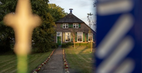 Een traditioneel Nederlands huis met een rieten dak, geflankeerd door bomen en bloemen, gezien door een hek.