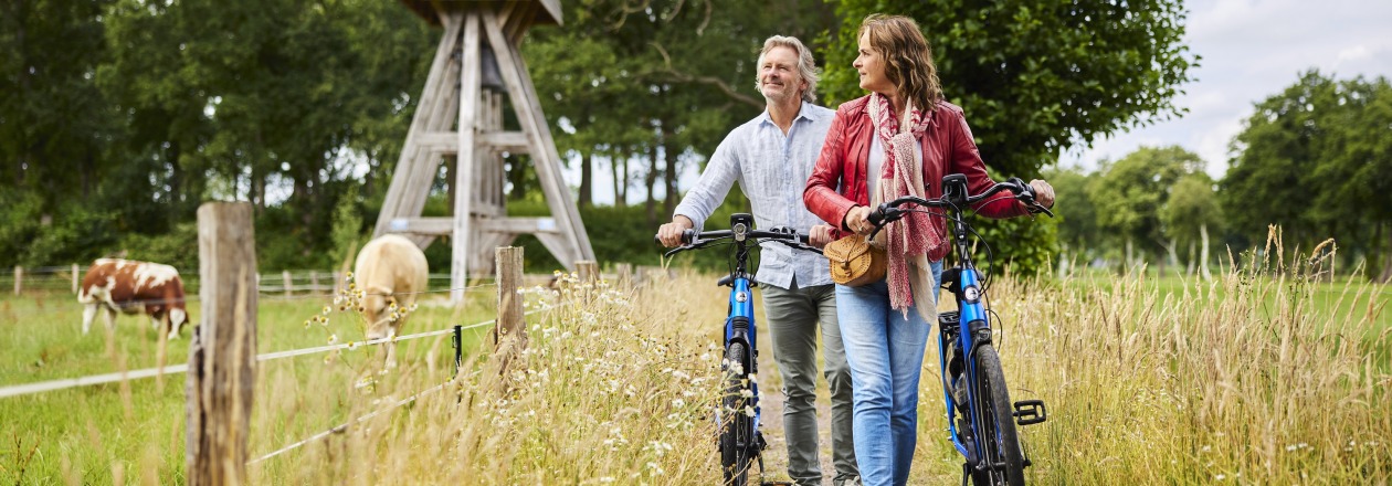 Een stel wandelt met fietsen over een landelijk pad langs een weiland met koeien en een houten klokkenstoel.