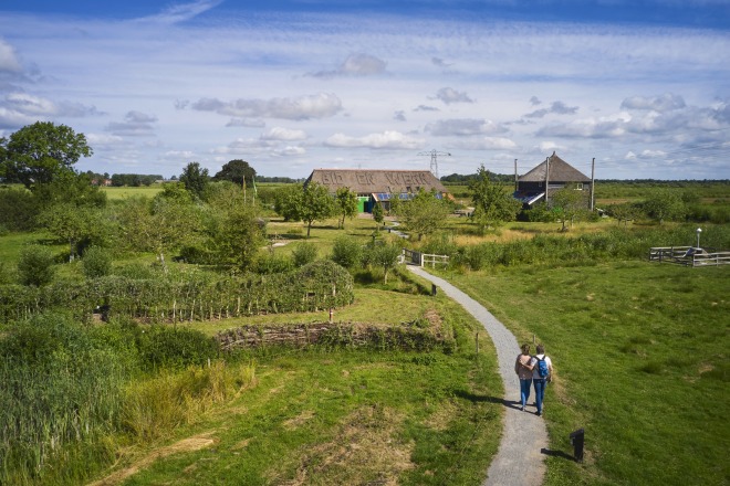 Twee mensen wandelen op een pad door een groen landschap met bomen en boerderijen onder een blauwe lucht.