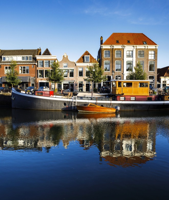 Historische grachtenpanden en woonboten weerspiegeld in het rustige water onder een heldere blauwe lucht.