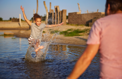 Een kind springt enthousiast in ondiep water, terwijl een volwassene toekijkt op een zonnige dag.
