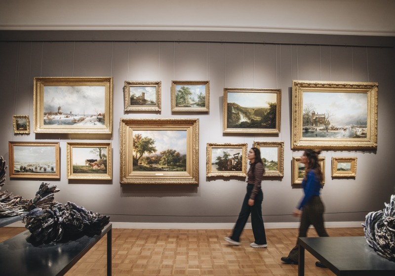 Twee vrouwen lopen langs een muur vol klassieke schilderijen in een kunstmuseum met houten vloer.