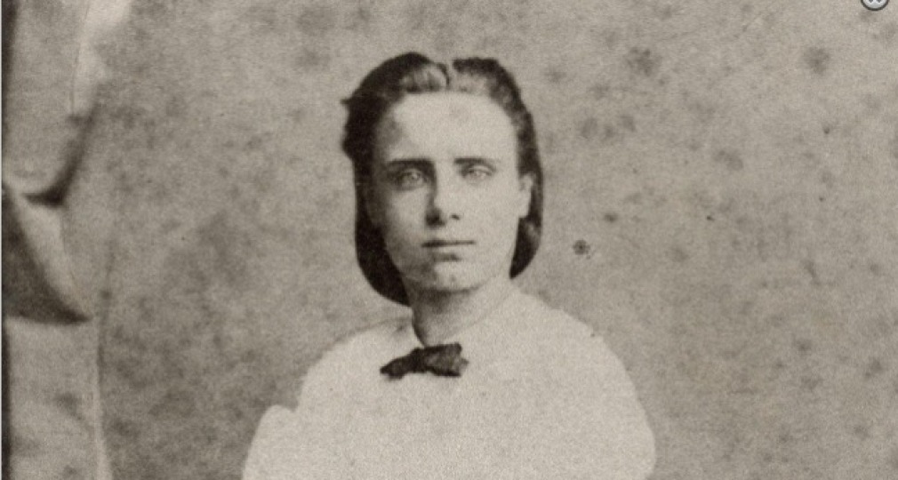 Zwart-witfoto van een jonge vrouw in een Victoriaanse jurk met een strikje, zittend en naar voren kijkend.