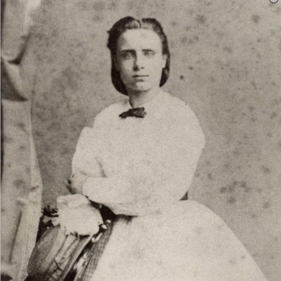 Zwart-witfoto van een jonge vrouw in een Victoriaanse jurk met een strikje, zittend en naar voren kijkend.