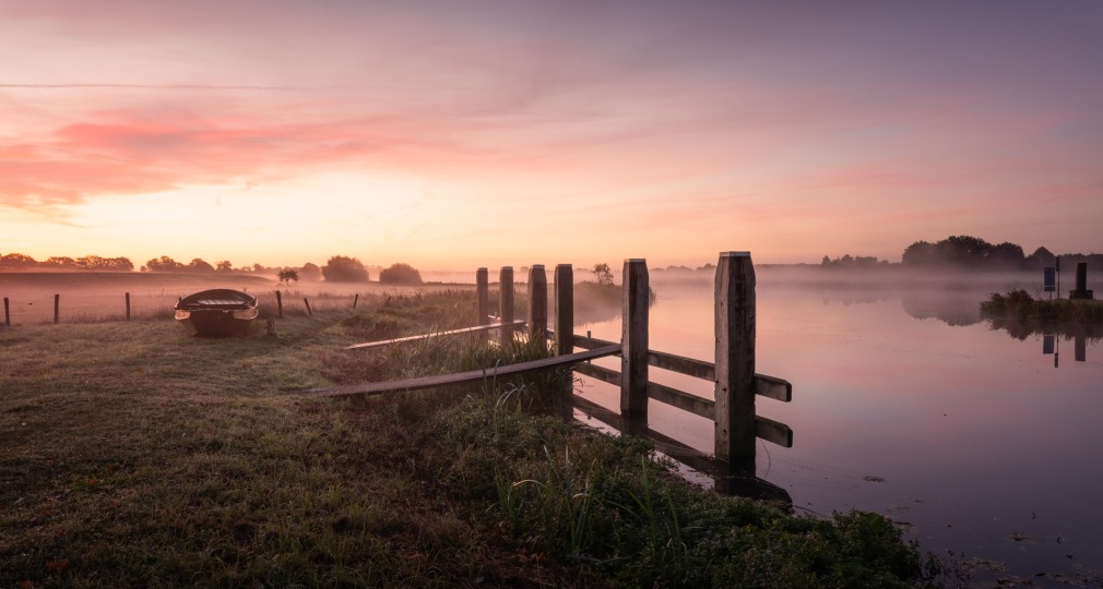 Een serene ochtendscène met een boot aan de oever, mist boven het water en een kleurrijke zonsopgang.