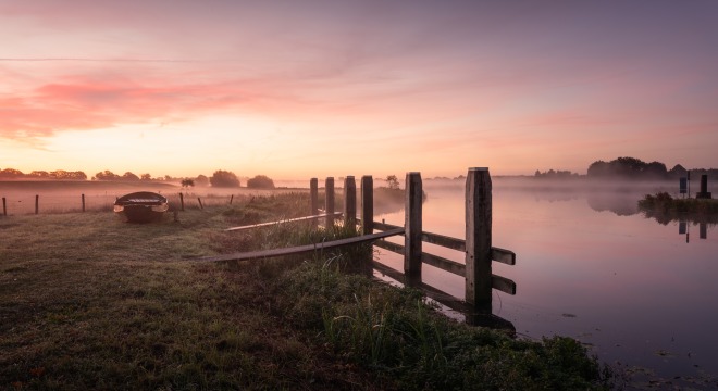 Een serene ochtendscène met een boot aan de oever, mist boven het water en een kleurrijke zonsopgang.