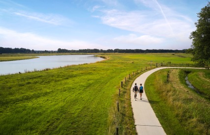 Twee mensen wandelen hand in hand op een kronkelend pad langs een rivier in een groen landschap.