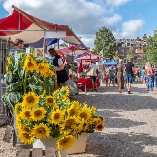 Mensen bezoeken een buitenmarkt met kraampjes, verse zonnebloemen en historische gebouwen op de achtergrond.