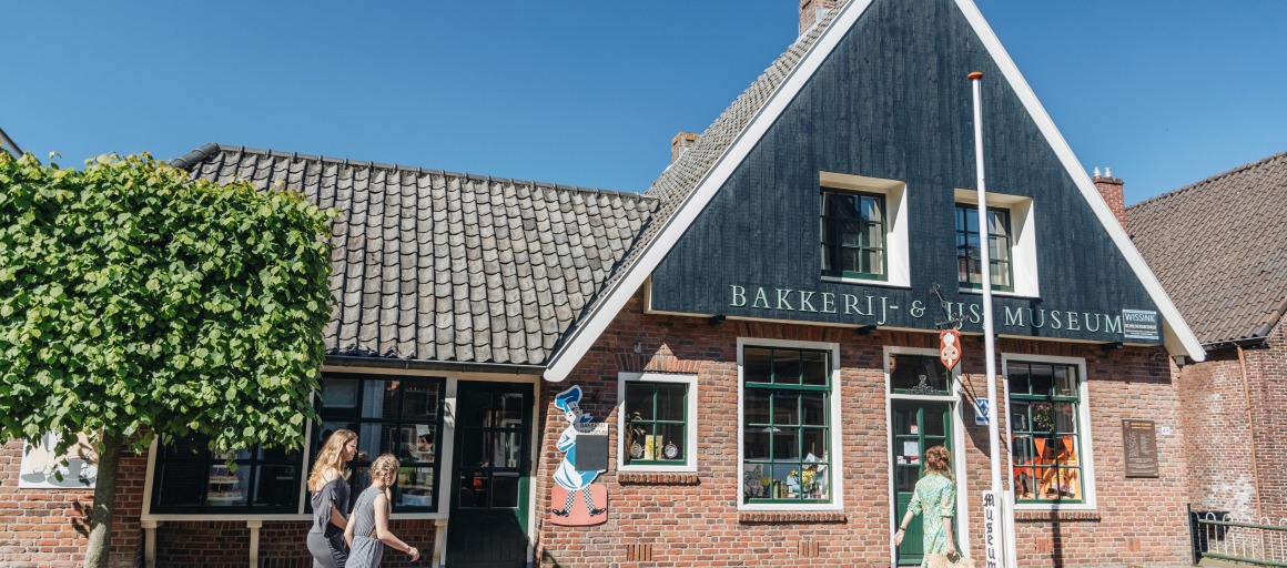 Twee vrouwen lopen langs het Bakkerij & Vis Museum in een karakteristiek Nederlands dorp.