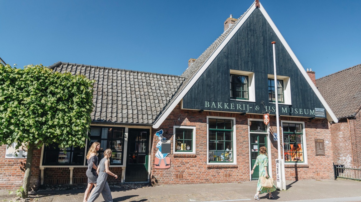 Twee vrouwen lopen langs het Bakkerij & Vis Museum in een karakteristiek Nederlands dorp.