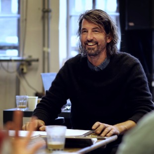 Man met halflang haar en baard lacht terwijl hij aan tafel zit in een moderne kantooromgeving.