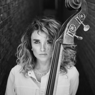 Zwart-wit portret van een vrouw met krullend haar die naast een contrabas tussen bakstenen muren staat.