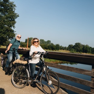 Oud stel fietst samen over een houten brug met uitzicht op een rivier en groene bomen op een zonnige dag.